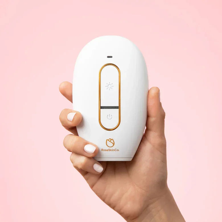 Hand holding RoseSkinCo Lumi IPL hair removal handset, white and gold, on pink background